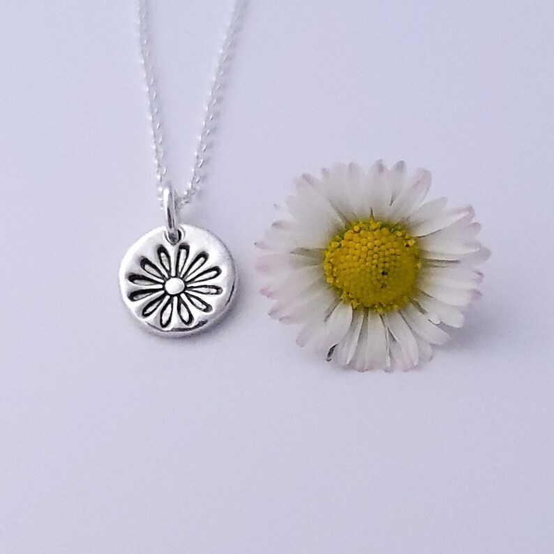 May include: A silver necklace with a small, round pendant featuring a daisy design. The pendant is textured and has a raised center. A white daisy with a yellow center is shown next to the necklace.