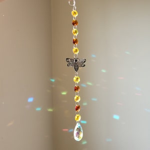 May include: A crystal suncatcher with a bee charm and yellow and amber beads hanging from a silver chain. The suncatcher has a teardrop-shaped crystal at the bottom.