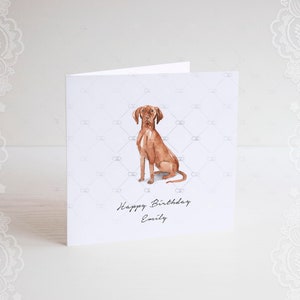 Pode incluir: Um cart&atilde;o branco com uma ilustra&ccedil;&atilde;o em aquarela de um c&atilde;o marrom sentado. O cart&atilde;o tem o texto "Happy Birthday Emily" impresso nele.