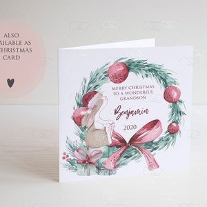 May include: A Christmas card featuring a watercolor illustration of a bunny wearing a Santa hat, sitting on a gift box with a red bow. The bunny is surrounded by a green wreath with red berries and a pink bow. The text "Merry Christmas to a wonderful Grandson Benjamin 2020" is written on the card.