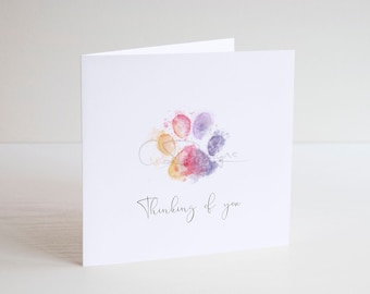 May include: A white card with a watercolor paw print in shades of orange, pink, purple, and blue. The text "Thinking of you" is written in black cursive below the paw print.