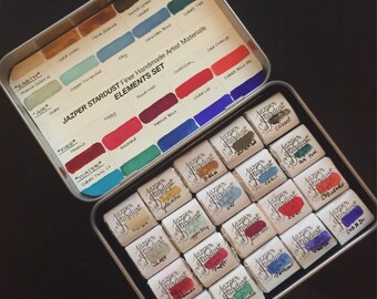 May include: A metal tin with a set of 24 watercolor paints. The tin has a label that says "Jazper Stardust Finer Handmade Artist Materials Elements Set". The paints are arranged in a grid pattern and are labeled with their names, such as "Gold Ochre", "Cassel", and "Emerald".