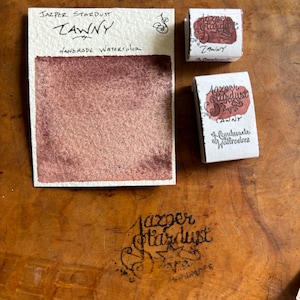 May include: A watercolor paint pan labeled "Jasper Stardust Tawny Handmade Watercolor". The pan is filled with a dark reddish-brown watercolor paint.