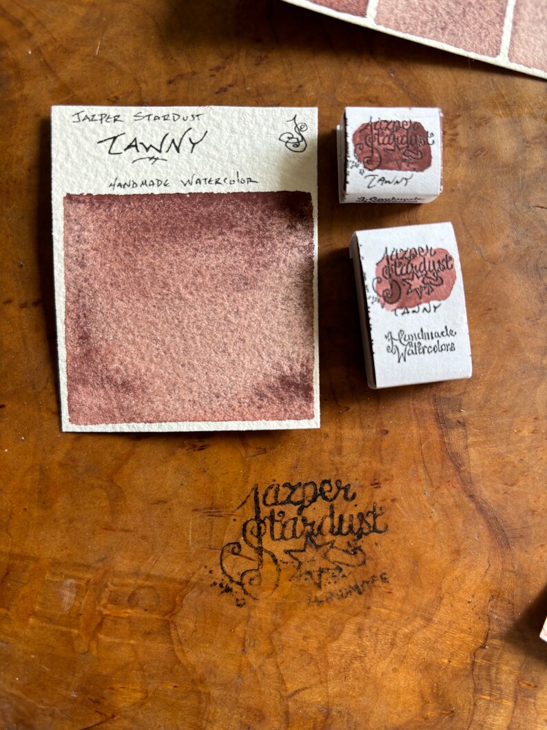May include: A watercolor paint pan labeled "Jasper Stardust Tawny Handmade Watercolor". The pan is filled with a dark reddish-brown watercolor paint.