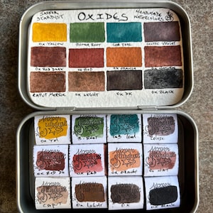 May include: A metal tin of watercolor paints labeled "Jazpar Stardust Oxides Handmade Watercolors". The tin contains 12 watercolor pans, each labeled with the color name and "Jazpar Stardust". The colors include: Ox Yellow, Bitter Root, Cob Teal, Celtic Violet, Ox Red Dark, Ox Red, Ox Orange, Da Natural, Caput Mortuum, Ox Light, Ox DK, and Ox Black.