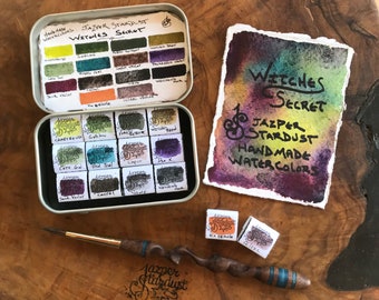 May include: A set of 18 watercolor paints in a tin box. The paints are labeled with names like "Chartreuse", "Cote Gold", and "Wendily Dark". The box also includes a small brush and a card with the words "Witches Secret Jazper Stardust Handmade Watercolors".