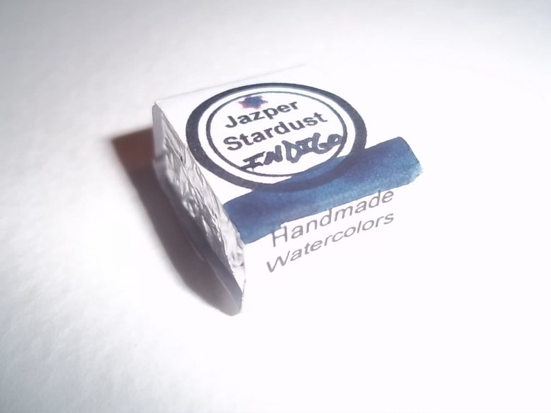 May include: A small, square, white container of handmade watercolor paint. The container has a black and white label with the text "Jazper Stardust Indigo" and "Handmade Watercolors".