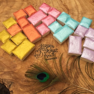 May include: A set of 18 watercolor paint pans in various metallic colors, including gold, silver, pink, orange, and blue. The pans are arranged in a semi-circle on a wooden surface. The text "Jazper Stardust Handmade" is visible in the center of the image.