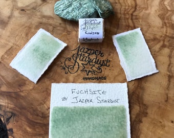 May include: A small container of green watercolor paint labeled "Fuchsite by Jazper Stardust Handmade".  Two swatches of the paint are shown on white paper.