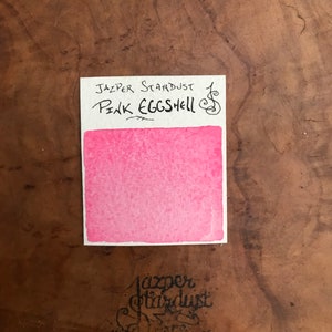 May include: A pink watercolor swatch with the text "Jasper Stardust Pink Eggshell" above it. The swatch is on a brown wooden surface.