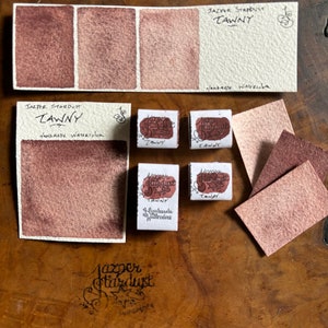 May include: Three watercolor paint swatches in shades of brown, labeled "Jasper Stardust Tawny" and "Handmade Watercolor".  There are also four small watercolor pans labeled "Jasper Stardust Tawny" and "Handmade Watercolor".