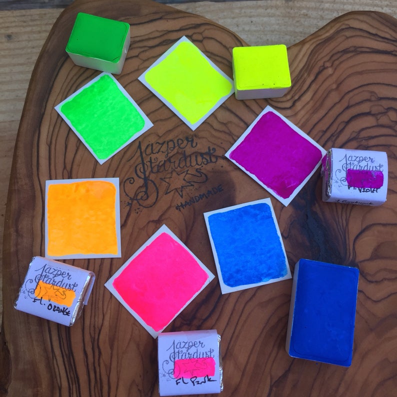 May include: Assortment of eight watercolor paint pans in various neon colors, including yellow, green, orange, pink, blue, and purple. The pans are labeled "Jasper Stardust" and "Handmade".
