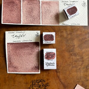 May include: Three watercolor paint swatches in shades of brown, labeled "Jazper Stardust Tawny" and "Handmade Watercolor".  The paint is in a small, square pan with a label that reads "Jazper Stardust Tawny" and "Handmade Watercolors".