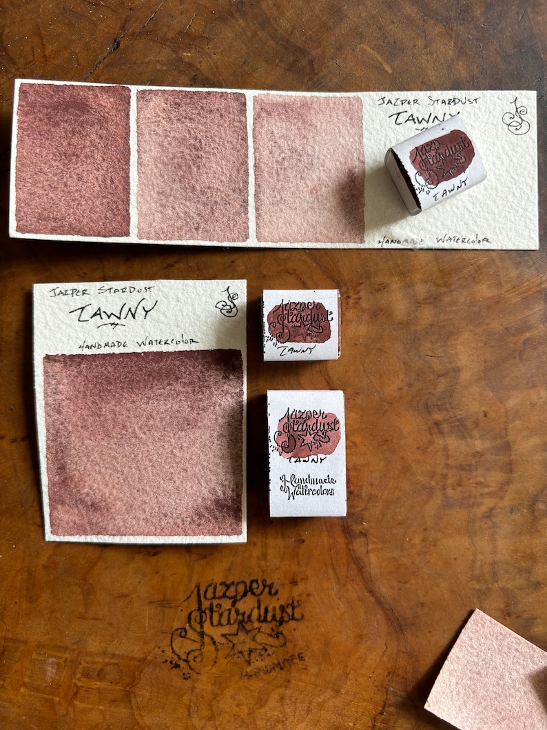 May include: Three watercolor paint swatches in shades of brown, labeled "Jazper Stardust Tawny" and "Handmade Watercolor".  The paint is in a small, square pan with a label that reads "Jazper Stardust Tawny" and "Handmade Watercolors".