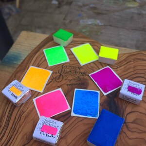 May include: A collection of watercolor paint pans in various bright colors, including yellow, green, pink, blue, and purple. The pans are labeled "Jazper Stardust Handmade".