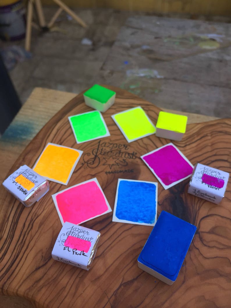 May include: A collection of watercolor paint pans in various bright colors, including yellow, green, pink, blue, and purple. The pans are labeled "Jazper Stardust Handmade".
