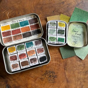 May include: Two open metal tins of watercolor paints. The top tin contains 12 watercolor paints labeled "Oxides" and the bottom tin contains 12 watercolor paints labeled "Jazper Stardust". The tin on the right contains 6 watercolor paints labeled "Green Forest Extender".