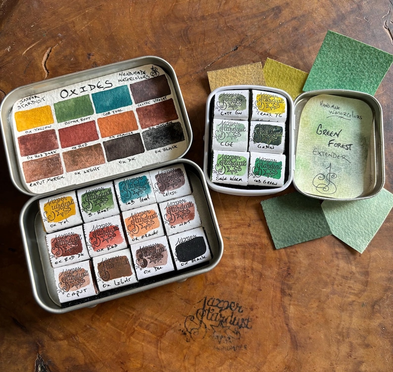May include: Two open metal tins of watercolor paints. The top tin contains 12 watercolor paints labeled "Oxides" and the bottom tin contains 12 watercolor paints labeled "Jazper Stardust". The tin on the right contains 6 watercolor paints labeled "Green Forest Extender".