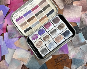 May include: A tin of watercolor paints called "Midnight Mystery" by Jazper Stardust. The tin contains 16 watercolor pans in a variety of colors, including blues, greens, browns, and purples. The pans are arranged in a grid pattern and are labeled with their names. The tin is surrounded by small squares of paper that have been painted with the watercolors.