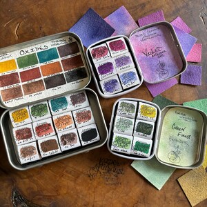 May include: Four metal tins of watercolor paints. The tins are labeled "Oxides Watercolor", "Violet Extender Set", "Green Forest Extender", and "Hand-mixed Watercolors". Each tin contains a set of watercolor paints in different colors.