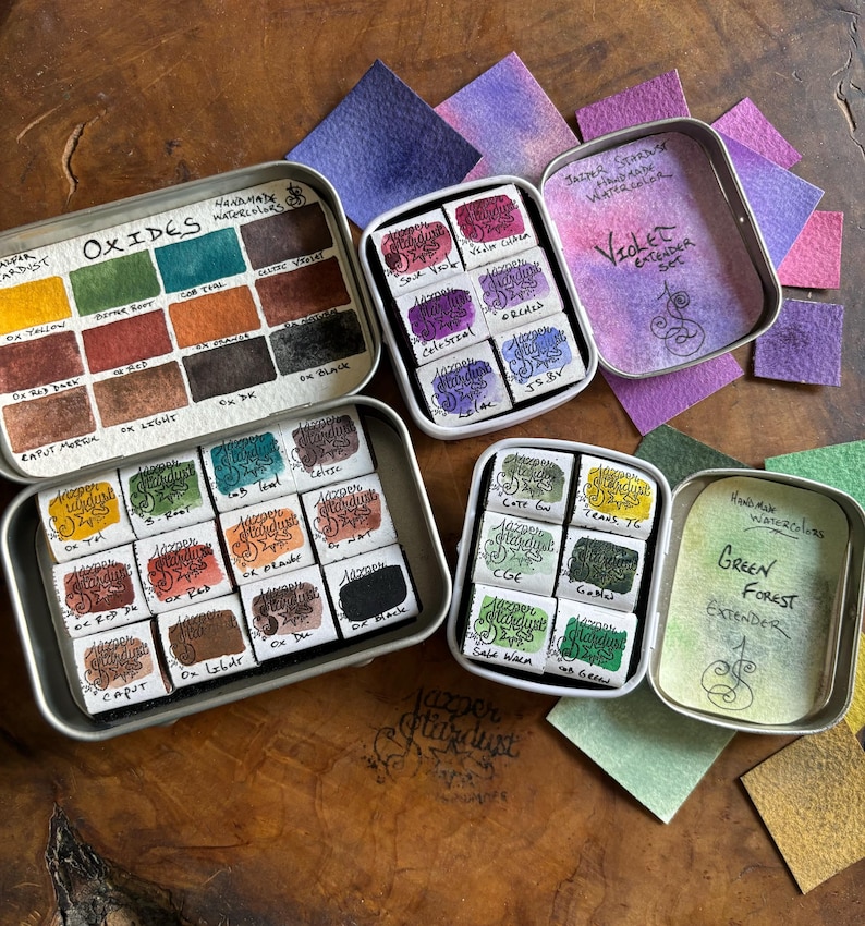 May include: Four metal tins of watercolor paints. The tins are labeled "Oxides Watercolor", "Violet Extender Set", "Green Forest Extender", and "Hand-mixed Watercolors". Each tin contains a set of watercolor paints in different colors.