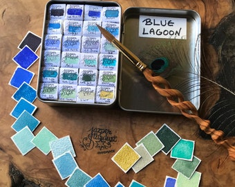 May include: A watercolor paint set with a wooden handled brush and a peacock feather. The set includes a metal tin labeled "Blue Lagoon" and a variety of square watercolor swatches in shades of blue, green, and yellow.