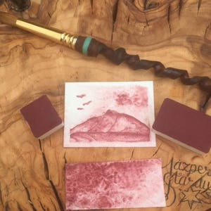 Op de afbeelding: Een close-up van een rode aquarelverfset met een penseel en een vel papier met een aquarel schilderij van een bergketen. Het papier ligt op een houten oppervlak.