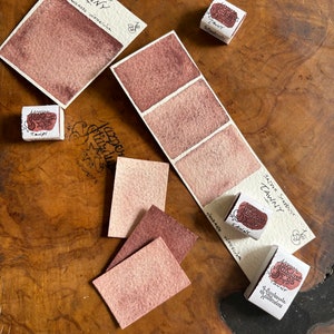 May include: Two watercolor paint pans labeled "Jazper Stardust Tawny" with a sample of the color on a white card. The color is a light brown with a reddish hue. The paint pans are on a wooden surface.