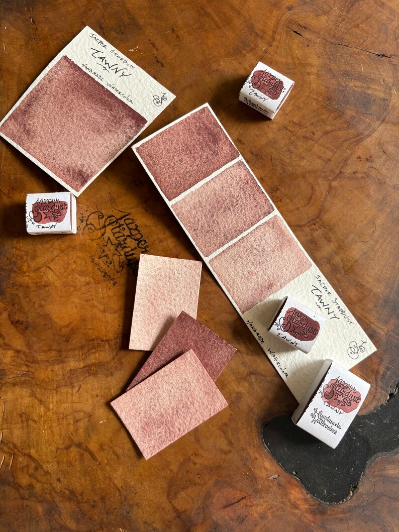 May include: Two watercolor paint pans labeled "Jazper Stardust Tawny" with a sample of the color on a white card. The color is a light brown with a reddish hue. The paint pans are on a wooden surface.