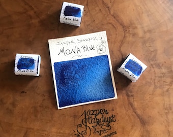 May include: A watercolor paint swatch labeled "Mona Blue" with a deep blue color gradient. Three small wrapped paint cubes with "Jasper Stardust Mona Blue" text are scattered around the swatch. The Jasper Stardust logo and "handmade" text are also visible.