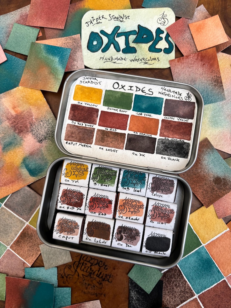 May include: A metal tin of handmade watercolor paints in a variety of earthy tones. The tin is labeled "Jasper Stardust Oxides Handmade Watercolors". The colors include Ox Yellow, Bitter Root, Cob Teal, Celtic Violet, Ox Red Dark, Ox Red, Ox Orange, Ox Light, Ox DK, Ox Black, Ox Tel, S-Root, Cob Teal, Celtic, Ox Red DK, Ox Red, Ox Pravee, Nat, Caput, Ox Lebur, Ox Du, and Ox Black.