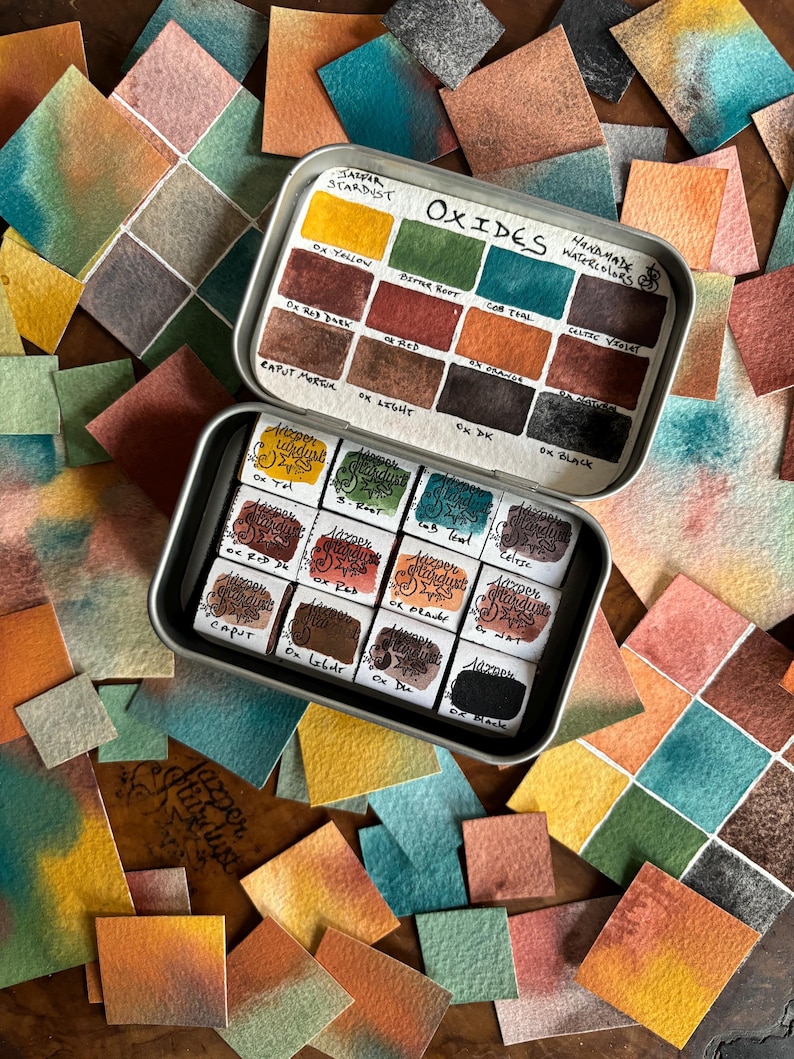 May include: A tin of handmade watercolor paints in a variety of earthy tones, including yellow, red, brown, green, and black. The tin is labeled "Oxides" and "Handmade Watercolors".