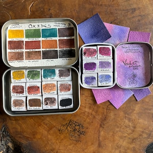 May include: A set of watercolor paints in a tin box. The set includes a variety of colors, including yellow, orange, red, brown, green, blue, and black. The tin box is labeled "Jazper Stardust" and "Oxides Handmade Watercolors".