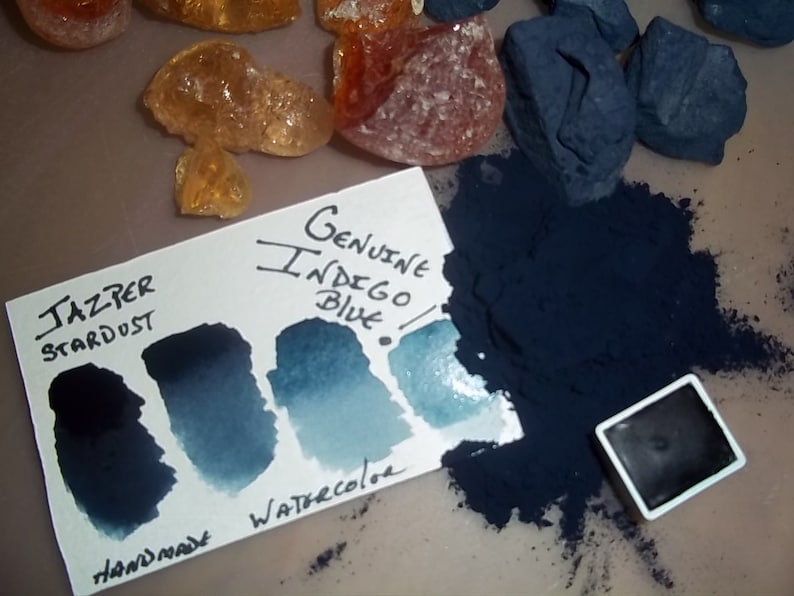 May include: A watercolor paint pan with a label that reads "Genuine Indigo Blue" and "Jasper Stardust" with a handmade label. The paint pan is surrounded by a pile of indigo blue powder and a few small rocks.