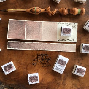 May include: A watercolor paint pan set with the brand name "Jazper Stardust Handmade" and the color "Celtic Violet". The paint pan is a light purple color and is shown next to a swatch of the color on a white paper.
