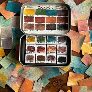 May include: A metal tin of watercolor paints labeled "Oxides" with the brand name "Jasper Stardust" and "Handmade Watercolors". The tin contains 12 small pans of watercolor paint in various shades of brown, red, orange, yellow, green, and black. The paint pans are labeled with the color name and the word "Ox" for oxide.