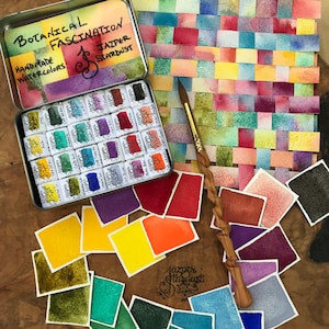 May include: A set of 24 watercolor paints in a tin box. The paints are arranged in a grid pattern and are labeled with the name of the color. The box is labeled "Botanical Fascination Handmade Watercolors Jazper Stardust".