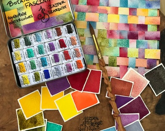 May include: A set of 24 watercolor paints in a tin box. The paints are arranged in a grid pattern and are labeled with the name of the color. The box is labeled "Botanical Fascination Handmade Watercolors Jazper Stardust".