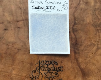 Può includere: Un piccolo cartoncino bianco con il testo "Jazper Stardust" e "Sodalite Gen." scritto in inchiostro nero. Il cartoncino è su una superficie di legno.