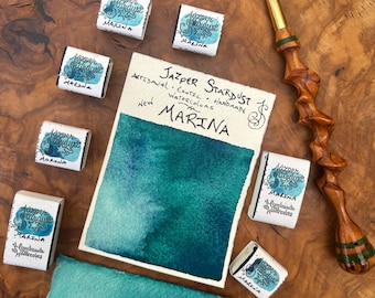 Può includere: Una collezione di sei piccoli godet quadrati di acquerello etichettati Jazper Stardust Marina, un campione di carta rettangolare con un lavaggio ad acquerello verde acqua e un pennello in legno con manico intrecciato. La carta riporta il testo Jazper Stardust Artisanal Exotic Handmade Watercolors New Marina.