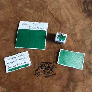 May include: A watercolor paint sample in a small tin labeled "Cobalt Green by Jazper Stardust" with the words "Lightfast? Excellent, Opacity? Opaque, Granular? Smooth" written on a white card. The paint is a vibrant green color.