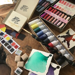 May include: A set of watercolor paints in a variety of colors, including red, green, blue, and black. The paints are in small bottles and are arranged in a box. There are also some watercolor pencils and a paintbrush in the image.