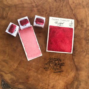 Pode incluir: Três pequenas bandejas de tinta aquarela etiquetadas como "Jazper Stardust Ruby" e uma amostra de tinta aquarela maior mostrando a cor "Ruby" com o texto "Jazper Stardust Ruby" acima.