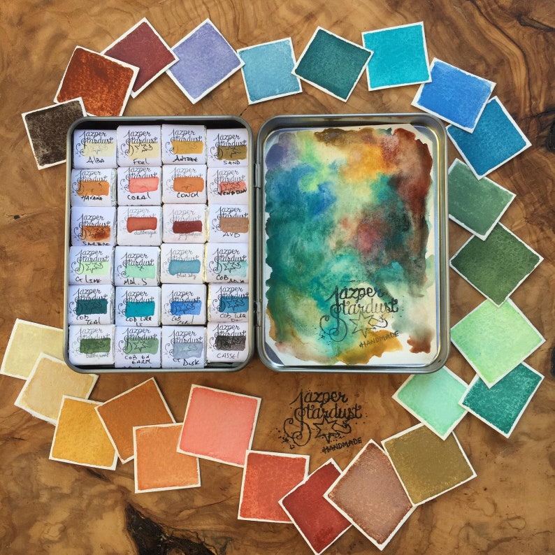 May include: A metal tin filled with 36 watercolor paint pans. The pans are arranged in a grid pattern and labeled with names like "Alaa", "Coral", "Cob", and "Dusk". The tin is open and the paint pans are visible. The tin is surrounded by small squares of watercolor paper, each painted with a different color from the paint pans. The text "Jazper Stardust Handmade" is visible on the tin and on one of the squares of watercolor paper.