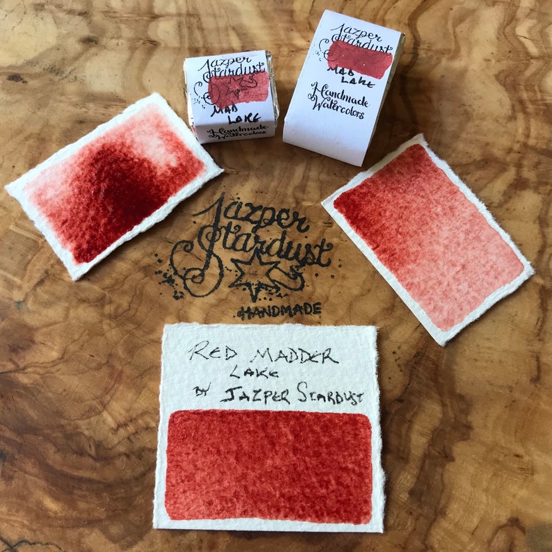 May include: A watercolor paint pan labeled "Red Madder Lake" by Jazper Stardust. The pan is handmade and comes in a small, square container. The paint is a deep red color.