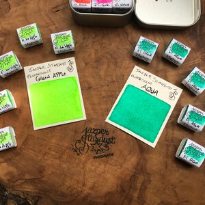 May include: Two watercolor paint pans, one green apple and one aqua, with the brand name Jazper Stardust and the words "Fluorescent" and "Handmade".