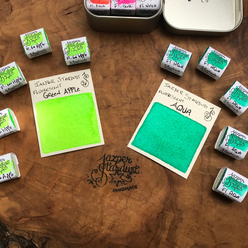 May include: Two watercolor paint pans, one green apple and one aqua, with the brand name Jazper Stardust and the words "Fluorescent" and "Handmade".