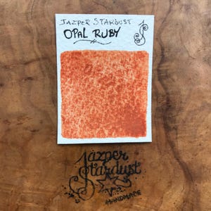 Könnte beinhalten: Eine kleine quadratische Probe der Aquarellfarbe "Opal Ruby" von Jazper Stardust. Die Farbe ist ein leuchtendes Orange-Rot mit einer gesprenkelten Textur.