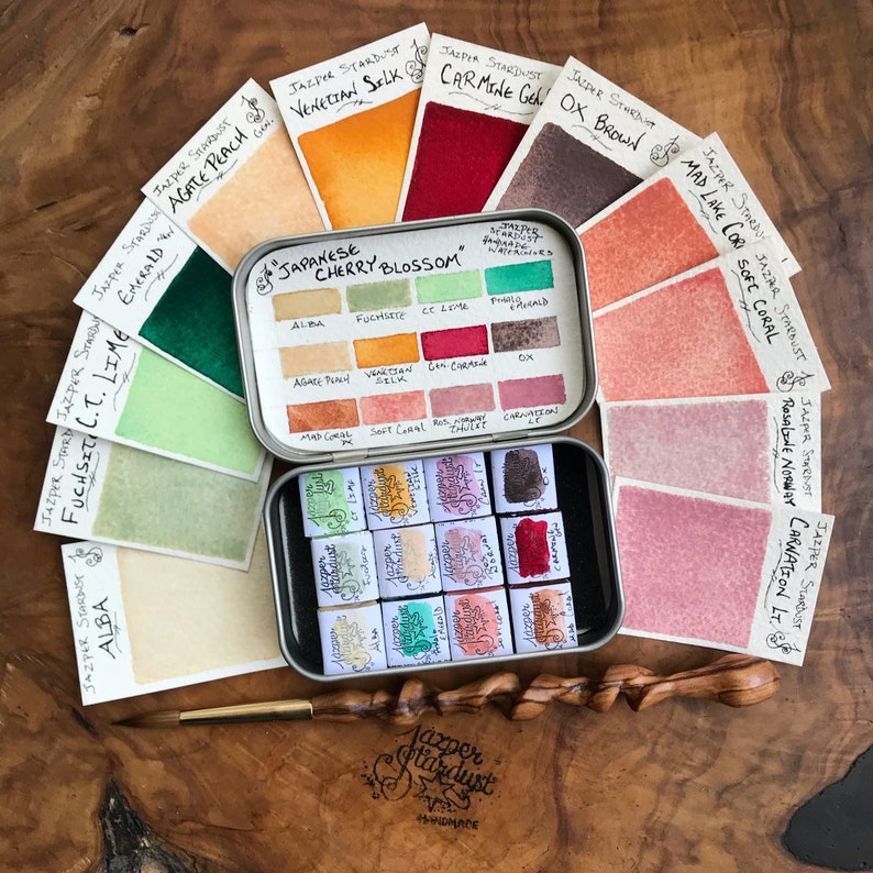 May include: A collection of watercolor paints in a variety of colors, including pinks, oranges, greens, and browns. The set includes a brush and two metal tins with the text "Japanese Cherry Blossom" and "Jasper Stardust Handmade".