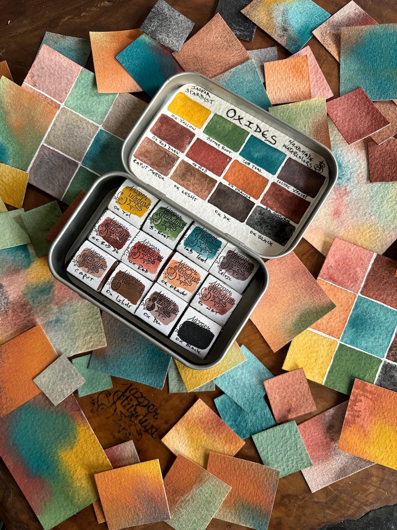 May include: A metal tin of watercolor paints with a variety of earthy tones. The tin is labeled "Oxides" and "Jatfar Stardust Watercolor". The paint colors include "Ox Yellow", "Ox Red", "Ox Bright", "Ox DK", "Ox Black", "Caput Morcea", "Corthal", "Caltec Viola", and more.